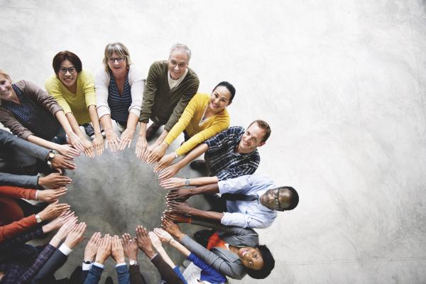 Image of fifteen diverse people standing around a round table with their arms extended, creating a circle with their hands